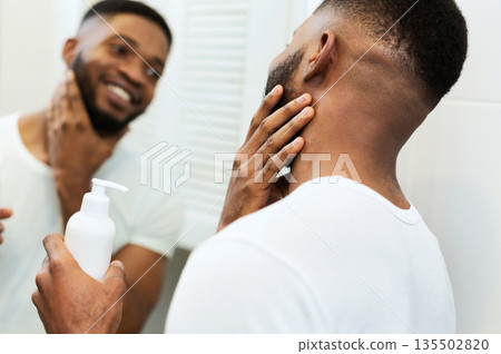 Facial care. Young black man applying moisturizer on beard in bathroom, looking in mirror Facial care. Young black man applying moisturizer on beard in bathroom, looking in mirror 135502820