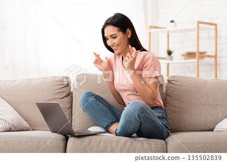 Cherished Wish. Portrait of young woman pleading for something with crossed fingers, using laptop sitting on couch Cherished Wish. Portrait of young woman pleading for something with crossed fingers, using laptop sitting on couch 135502839