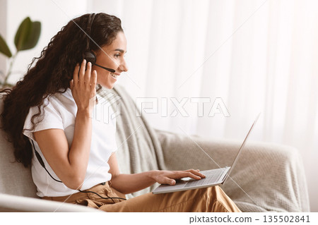 Side vire of cheerful pretty woman with dark curly hair having video conference from home, using headset and laprop while sitting on couch at living room, talking to friends or relatives, copy space Side vire of cheerful pretty woman with dark curly hair having video conference from home, using headset and laprop while sitting on couch at living room, talking to friends or relatives, copy space 135502841