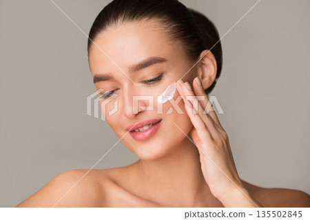 Portrait of woman applying moisturizer cream on face, standing over grey background 135502845