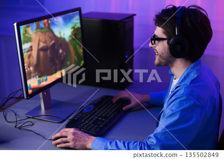 Male gamer playing shooter game on computer, wearing headphones and glasses 135502849