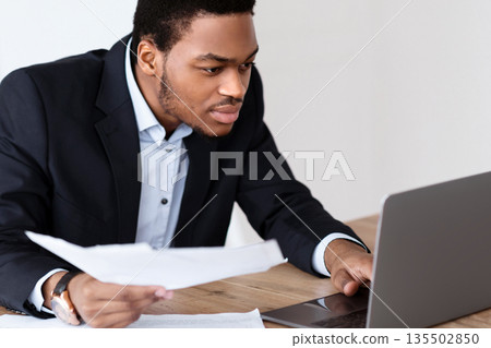 African american manager with documents checking marketing report, using laptop in office, copy space. Concentrated black businessman doing paperwork, checking data on computer 135502850