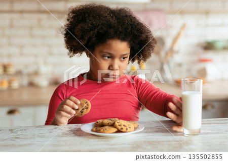 Grumpy Little Black Girl Refusing Milk And Eating Cookies In Kitchen, African Female Kid Choosing Fresh Baked Chocolate Biscuits Instead Of Healthy Calcium Drink While Having A Bite At Home, Closeup Grumpy Little Black Girl Refusing Milk And Eating Cookies In Kitchen, African Female Kid Choosing Fresh Baked Chocolate Biscuits Instead Of Healthy Calcium Drink While Having A Bite At Home, Closeup 135502855