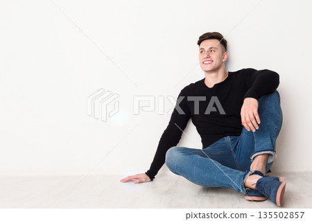 Smiling young man looks up, sitting on the floor at white background, copy space Smiling young man looks up, sitting on the floor at white background, copy space 135502857