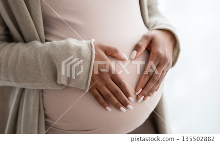Pregnant black woman holding palms in heart shape on her big tummy, cropped. Unrecognizable african american expecting lady standing by window at home. Pregnancy and maternity concept 135502882