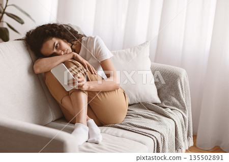 Lonely woman with smartphone sitting on sofa at home, waiting for message from lover, copy space. Upset young lady looking at mobile phone screen, having depression, living room interior 135502891
