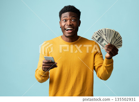 Emotional handsome african american young man in yellow using modern cellphone and showing bunch of cash on blue studio background, recommending newest mobile application, copy space Emotional handsome african american young man in yellow using modern cellphone and showing bunch of cash on blue studio background, recommending newest mobile application, copy space 135502931
