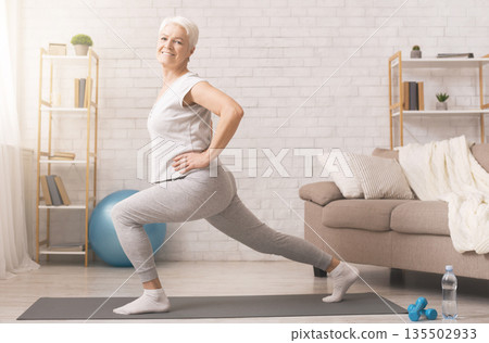 Legs stretching. Sporty senior lady doing lunges at home, empty space Legs stretching. Sporty senior lady doing lunges at home, empty space 135502933