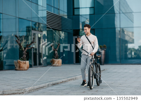 Surprised news and good offer. Shocked cheerful millennial male hipster manager in stylish clothes reads amazing message in phone, stands with bicycle near modern city office on street, outdoor 135502949
