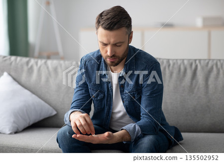Upset middle-aged bearded mad sitting on couch in living room and looking at pills in his hands, suffering from depression or having sexual dysfunction, copy space. Medication, medicine, pills Upset middle-aged bearded mad sitting on couch in living room and looking at pills in his hands, suffering from depression or having sexual dysfunction, copy space. Medication, medicine, pills 135502952