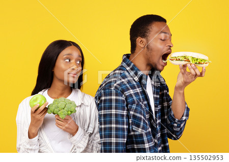 Healthy nutrition and fast food, diet and relationship. Funny millennial african american guy eats sandwich, wife with vegetables and fruits sadly looks at husband isolated on yellow background Healthy nutrition and fast food, diet and relationship. Funny millennial african american guy eats sandwich, wife with vegetables and fruits sadly looks at husband isolated on yellow background 135502953
