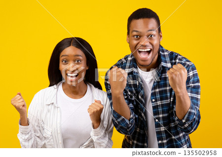 Human emotions and victory in online game. Surprised successful young african american male and female laugh and keep fists celebrating win, isolated on yellow background, studio shot, copy space 135502958