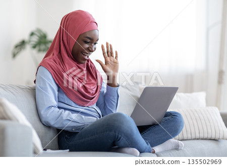 Cheerful black muslim lady in hijab making video call on laptop at home, waving at screen, chatting with family or friends, enjoying online communication, sitting on couch in living room, copy space 135502969