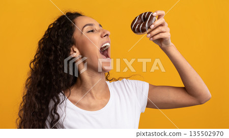Cheat Meal. Funny Hungry Latin Lady Holding Doughnut Eating Junk Food Posing With Wide Open Mouth Over Yellow Orange Studio Background. Unhealthy Nutrition And Binge Eating Habit Concept Cheat Meal. Funny Hungry Latin Lady Holding Doughnut Eating Junk Food Posing With Wide Open Mouth Over Yellow Orange Studio Background. Unhealthy Nutrition And Binge Eating Habit Concept 135502970