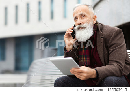 Elderly grey-haired man with long beard sitting on bench by office building, holding digital tablet and talking on mobile phone, having business call while resting on the street, looking at free space Elderly grey-haired man with long beard sitting on bench by office building, holding digital tablet and talking on mobile phone, having business call while resting on the street, looking at free space 135503045