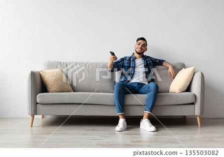 Portrait of Arab guy watching TV show or film, holding remote control, switching channels. Young smiling man spending weekend free time sitting on comfortable sofa at home in living room, copy space Portrait of Arab guy watching TV show or film, holding remote control, switching channels. Young smiling man spending weekend free time sitting on comfortable sofa at home in living room, copy space 135503082