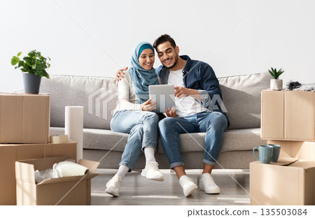 Joyful muslim family sitting on couch in new apartment, using pad, copy space. Happy middle-eastern young man and woman in hijab buying furniture for house, unpacking stuff after moving Joyful muslim family sitting on couch in new apartment, using pad, copy space. Happy middle-eastern young man and woman in hijab buying furniture for house, unpacking stuff after moving 135503084