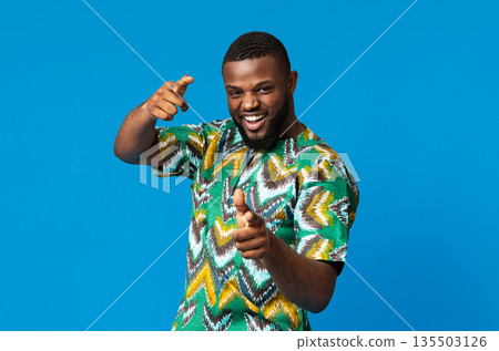 Playful black young man in colorful african costume, indicating to camera on blue studio background, empty space. Happy african american charismatic guy demonstrating traditional bright clothes Playful black young man in colorful african costume, indicating to camera on blue studio background, empty space. Happy african american charismatic guy demonstrating traditional bright clothes 135503126