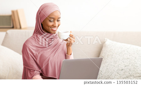 Online life concept. Black woman in hijab sitting on couch with laptop, drinking coffee, working or studying from home. Attractive African American lady in headscarf having business meeting on web Online life concept. Black woman in hijab sitting on couch with laptop, drinking coffee, working or studying from home. Attractive African American lady in headscarf having business meeting on web 135503152