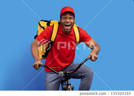 Fast Delivery Service. Overjoyed african american courier guy in red cap and uniform riding bicycle and wearing thermo backpack bag isolated on blue studio background. Shipment service during lockdown Fast Delivery Service. Overjoyed african american courier guy in red cap and uniform riding bicycle and wearing thermo backpack bag isolated on blue studio background. Shipment service during lockdown 135503183