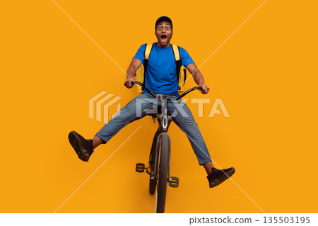 Full length of funny black delivery man in blue cap and uniform riding bicycle, wearing yellow thermo backpack bag isolated over bright orange blue studio background. Fast and quick shipment service 135503195