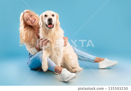 Love And Care Concept. Cheerful young lady patting her happy labrador spending time together, embracing pet, posing looking at camera sitting on the floor isolated on blue background, full body length 135503230