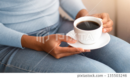 Coffee break. Cropped view of black woman holding cup of aromatic beverage while sitting on sofa indoors, closeup of hands. Unrecognizable African American lady enjoying her morning drink 135503246