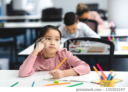Education And Learning Concept. Portrait of tired and bored small asian girl sitting at desk in classroom at school, writing in notebook and thinking, looking away at window, resting head on hand 135503247