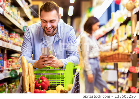 Smiling Male Customer Doing Grocery Shopping Using Smartphone Walking With Cart In Supermarket. Selective Focus. Man Using Groceries Shopping Application On Phone Bying Food In Super Market Smiling Male Customer Doing Grocery Shopping Using Smartphone Walking With Cart In Supermarket. Selective Focus. Man Using Groceries Shopping Application On Phone Bying Food In Super Market 135503250