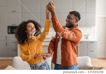 Cheerful millennial black spouses having fun together dancing to music in modern kitchen interior, smiling to each other. Couple having date or party. Relationship and romance concept 135503290