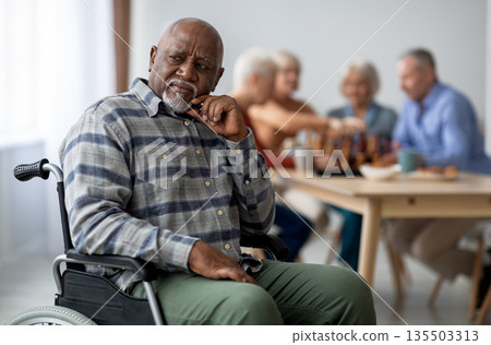 Disabled african american senior man in wheelchair feeling lonely, sitting aside of cheerful elderly men and women playing chess and chatting. Adaptation at retirement house concept Disabled african american senior man in wheelchair feeling lonely, sitting aside of cheerful elderly men and women playing chess and chatting. Adaptation at retirement house concept 135503313