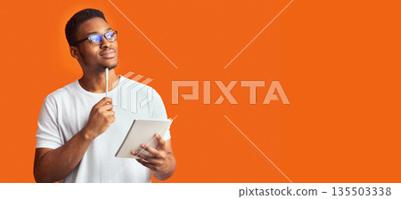 Pensive Redactor. Dreaming afro man in glasses taking notes, writing ideas, looking at copyspace, orange wall, panorama 135503338