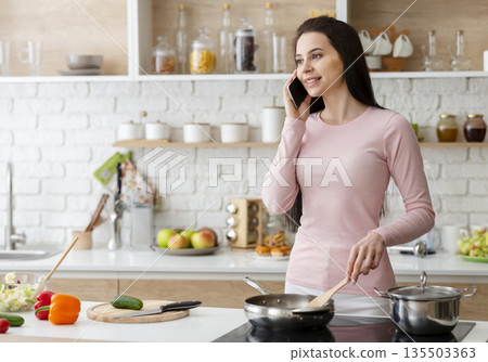 Female multitasking possibility. Young woman talking on phone and cooking lunch at kitchen, empty space Female multitasking possibility. Young woman talking on phone and cooking lunch at kitchen, empty space 135503363