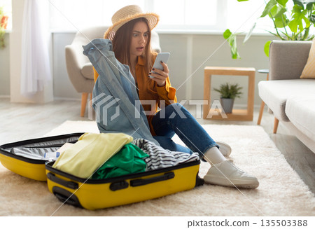 Terrified young woman reading shocking travel news or negative message about tourism on smartphone while packing suitcase at home. Cancelled flight, traveling problems during covid epidemic Terrified young woman reading shocking travel news or negative message about tourism on smartphone while packing suitcase at home. Cancelled flight, traveling problems during covid epidemic 135503388