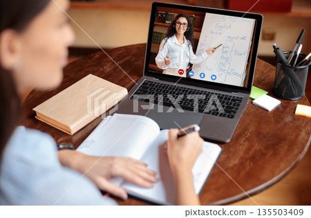 E-learning Concept. Over the shoulder back view of female student sitting at table, writing in notebook, taking notes, using laptop, having online class with English teacher. Distant studying E-learning Concept. Over the shoulder back view of female student sitting at table, writing in notebook, taking notes, using laptop, having online class with English teacher. Distant studying 135503409