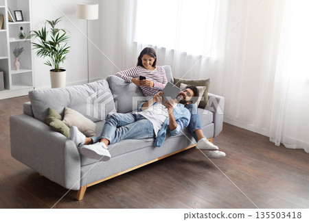High angle view of relaxed cheerful stylish millennial eastern lovers man and woman chilling on couch in cozy living room, using gadgets at home, scrolling on social media, reading online, copy space 135503418