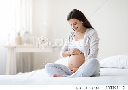 Happy pregnant woman sitting on bed in bedroom at home, hugging her big tummy. Happiness and feeling joyful as emotional well affect to baby neurological and psychological development, empty space Happy pregnant woman sitting on bed in bedroom at home, hugging her big tummy. Happiness and feeling joyful as emotional well affect to baby neurological and psychological development, empty space 135503442