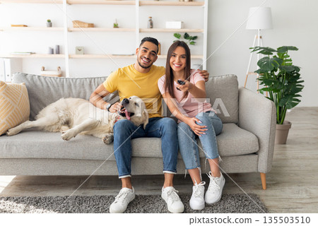 Young affectionate multiracial couple sitting on couch with pet dog and watching television. Diverse boyfriend and girlfriend spending time together, enjoying good movie at home Young affectionate multiracial couple sitting on couch with pet dog and watching television. Diverse boyfriend and girlfriend spending time together, enjoying good movie at home 135503510