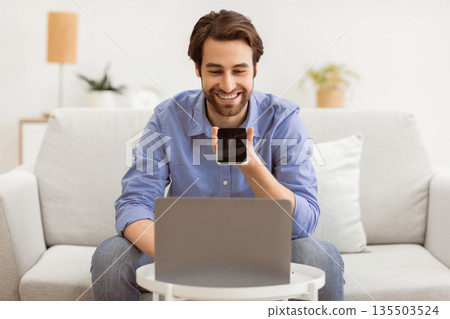 Cheerful Guy Using Voice Assistant On Mobile Phone Or Talking Using Loudspeaker Mode On Smartphone Sitting At Laptop Computer On Sofa At Home. Voice Search Application, Gadgets Lifestyle. Cheerful Guy Using Voice Assistant On Mobile Phone Or Talking Using Loudspeaker Mode On Smartphone Sitting At Laptop Computer On Sofa At Home. Voice Search Application, Gadgets Lifestyle. 135503524