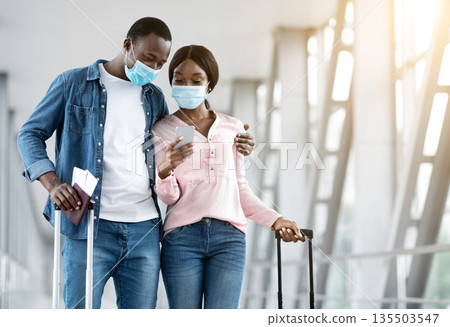 Checking Flight Information. Black couple wearing medical masks waiting for departure in airport, using smartphone together, copy space 135503547
