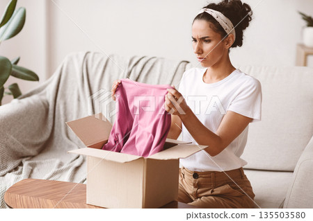 Disappointed young woman customer checking mail box, sitting on sofa at home, copy space. Lady opening delivery, checking clothes, having negative face expression. Delivery concept Disappointed young woman customer checking mail box, sitting on sofa at home, copy space. Lady opening delivery, checking clothes, having negative face expression. Delivery concept 135503580