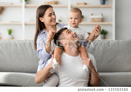 Happy Family Portrait. Cheerful young parents with little infant son having fun at home together, millennial mom and dad playing and laughing with cute toddler child in living room, copy space Happy Family Portrait. Cheerful young parents with little infant son having fun at home together, millennial mom and dad playing and laughing with cute toddler child in living room, copy space 135503588