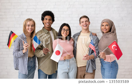 Student exchange, international friendship. Multiethnic young students with flags of different countries ready to study and look at camera on brick white wall background, studio shot, free space 135503589
