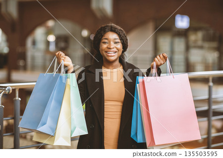 Best deal and huge discounts during Black Friday or final sale. Portrait of happy young african american woman shopper in coat shows many color shopping bags in city mall, copy space 135503595