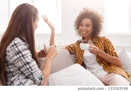 Two happy young female friends with coffee cups conversing in living room at home, chatting about their life and relations, gossip and slumber party concept, copy space 135503599