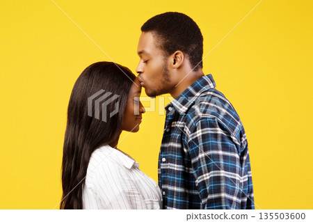 Romantic date, tenderness and relationship of young people. Calm african american male kisses on forehead wife and holds hands, isolated on yellow background, studio shot, profile, copy space Romantic date, tenderness and relationship of young people. Calm african american male kisses on forehead wife and holds hands, isolated on yellow background, studio shot, profile, copy space 135503600