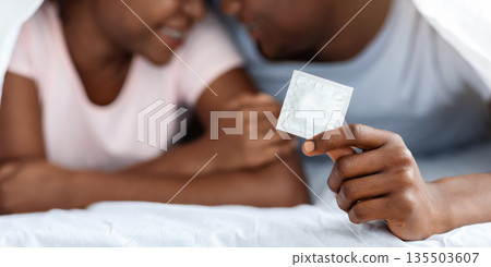 Contraceptive, contraception, safety sex concept. Selective focus on condom in black man hands cuddling with his woman under blanket at home. Closeup of condom in african american guy hand Contraceptive, contraception, safety sex concept. Selective focus on condom in black man hands cuddling with his woman under blanket at home. Closeup of condom in african american guy hand 135503607