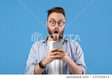 Shocked Caucasian guy staring at cellphone with open mouth over blue studio background, panorama. Surprised young man with mobile device learning unbelievable news, amazed at promo sale Shocked Caucasian guy staring at cellphone with open mouth over blue studio background, panorama. Surprised young man with mobile device learning unbelievable news, amazed at promo sale 135503613