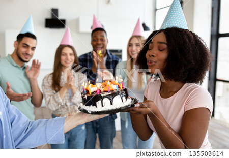 Beautiful black lady blowing candles on birthday cake, closing eyes, making wish, celebrating birthday with her diverse friends at home. Group of young people wearing party hats, having fun 135503614