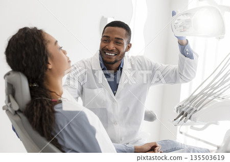 Cheerful black dentist turning on equipment before check up and greeting female patient Cheerful black dentist turning on equipment before check up and greeting female patient 135503619